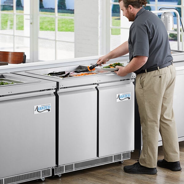 An Avantco stainless steel refrigerated salad bar on a counter in a commercial kitchen with a man preparing food.