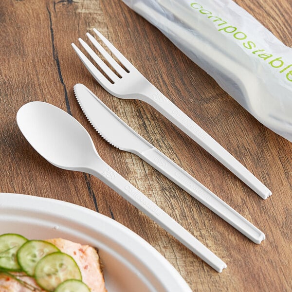 A wrapped EcoChoice white compostable CPLA fork and spoon next to a plate with a plastic fork.