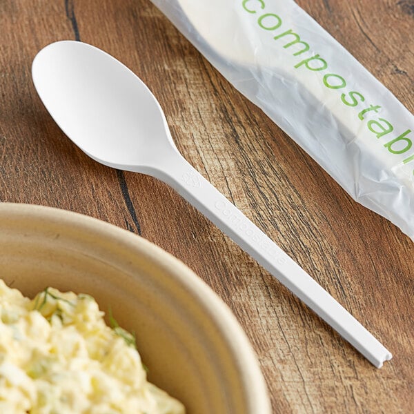 A bowl of food with a EcoChoice compostable white CPLA spoon next to a plastic bag of compostable materials.