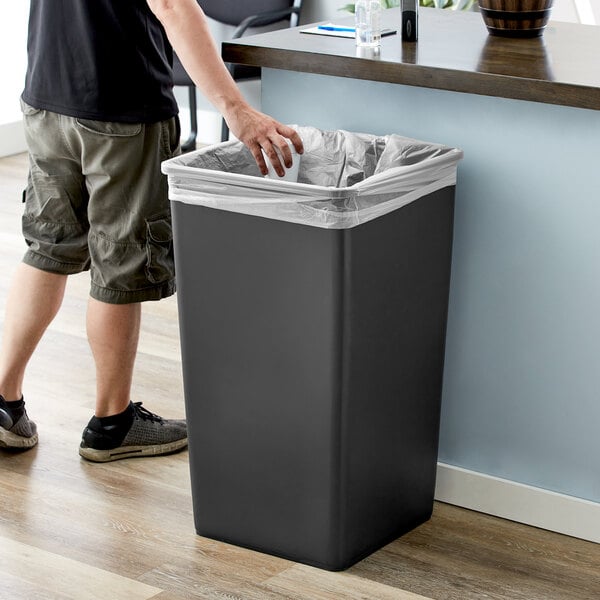 A man standing next to a black rectangular Lavex trash can with a plastic bag over it.