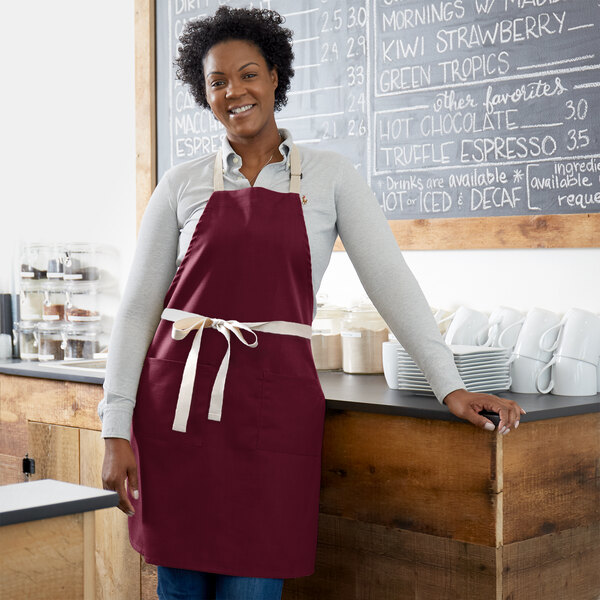 A woman wearing a Choice burgundy poly-cotton adjustable bib apron with natural webbing accents.