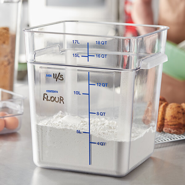 A person using a measuring cup to pour white powder into a clear Carlisle square food storage container.