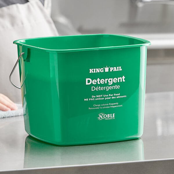 A person holding a green Noble Products detergent pail on a counter.