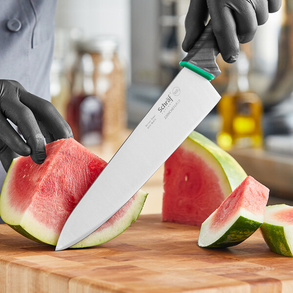 A person in gloves uses a Schraf chef knife with a green handle to cut a watermelon on a wooden surface.