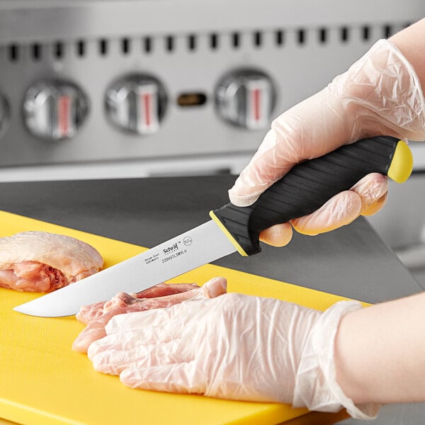 A person in gloves using a Schraf utility knife with a yellow and black TPRgrip handle to cut raw meat on a cutting board.