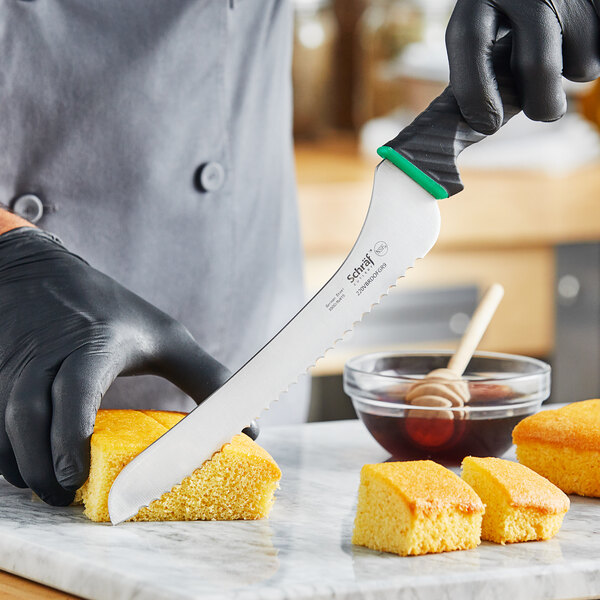 A person wearing a black glove uses a Schraf serrated bread knife to cut a piece of bread.
