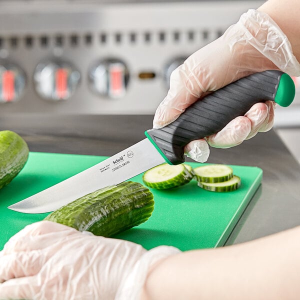 A person in gloves holding a Schraf utility knife with a green and black handle cutting a cucumber.