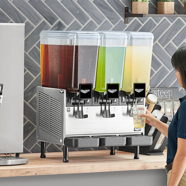 A woman using a Vollrath refrigerated beverage dispenser with three liquid dispensers.