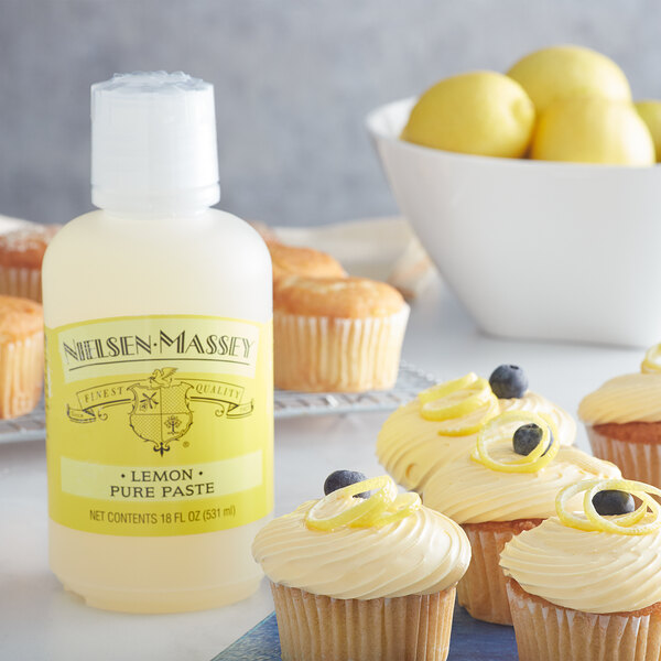 A cupcake with yellow frosting and a lemon peel on top next to a bottle of Nielsen-Massey Pure Lemon Paste.