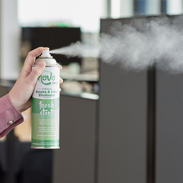 A hand holding a green and white Novo by Noble Chemical Fresh Start air freshener spray can.