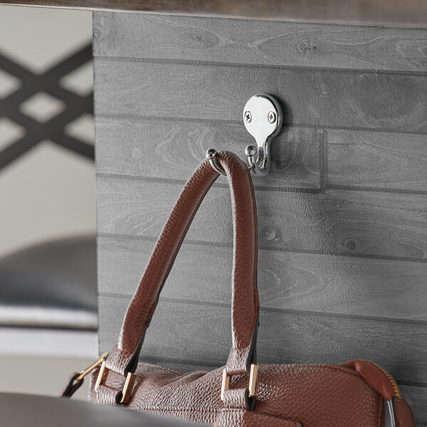 A brown leather purse hanging on a Lancaster Table & Seating double chrome bar purse hook on a table in a restaurant.