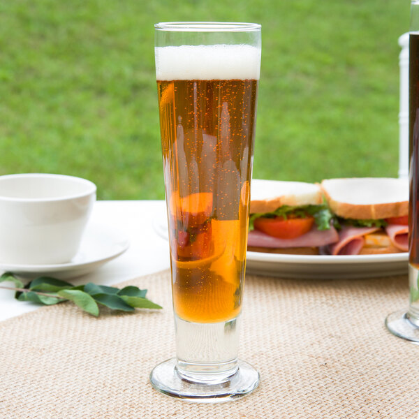 A Libbey tall footed pilsner glass of beer on a table next to sandwiches.