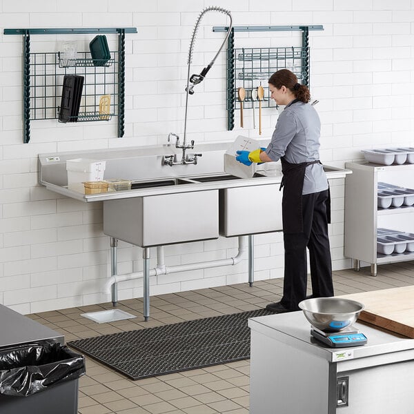 A woman cleaning a Regency stainless steel two compartment sink in a commercial kitchen.