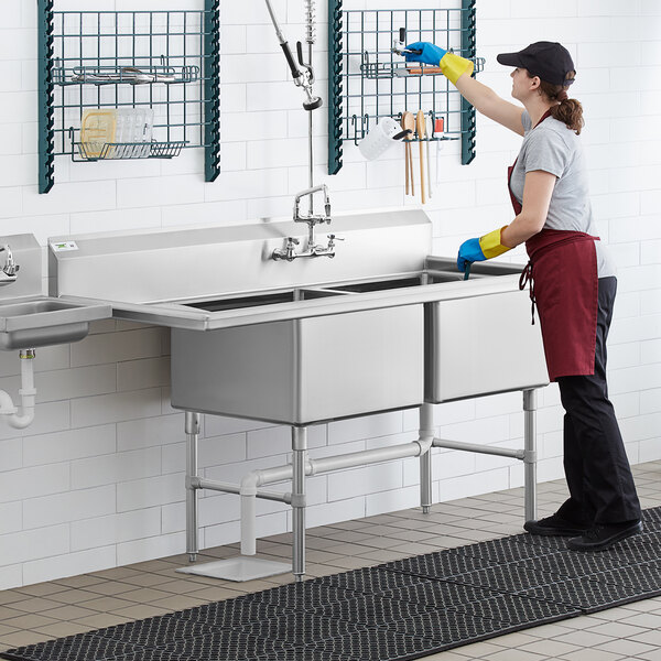 A woman in a black apron and gloves washing a Regency stainless steel two compartment sink on a counter.