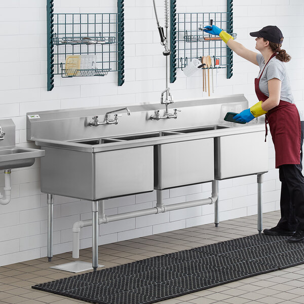 A woman wearing gloves cleaning a Regency stainless steel 3 compartment sink in a professional kitchen.