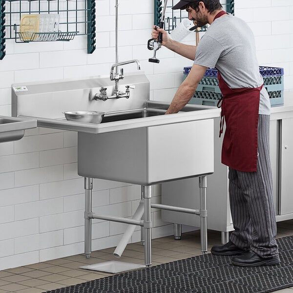 A man in a red apron washing his hands in a Regency stainless steel commercial sink with a left drainboard.