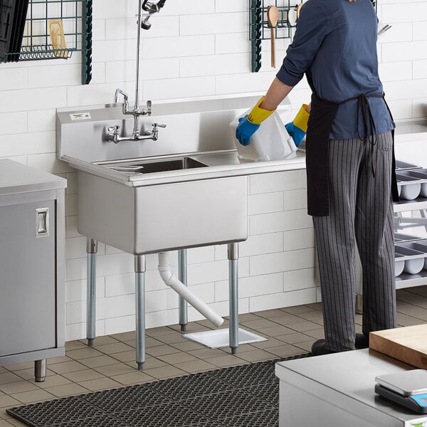 A person in a black apron and gloves washing dishes in a Regency stainless steel commercial sink with a right drainboard.