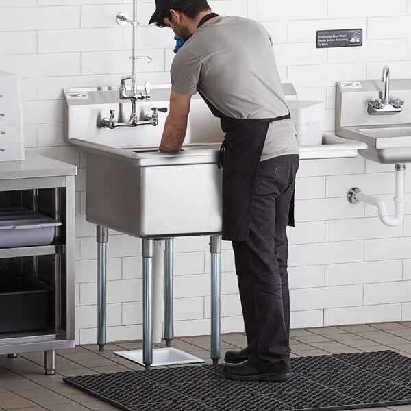 A man in a black shirt and apron standing at a Regency stainless steel commercial sink with a drainboard.