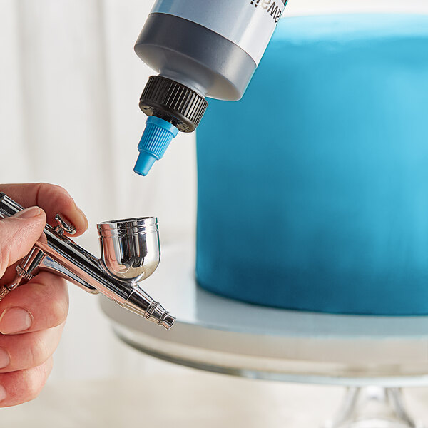 A hand using a silver airbrush to spray blue coloring onto a cake.