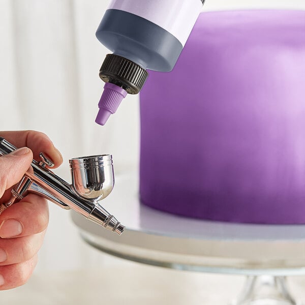A person using a Chefmaster airbrush to spray purple food coloring on a cake.