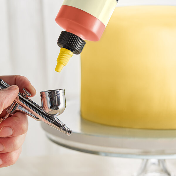 A finger holding a bottle of Chefmaster Canary Yellow airbrush color over a cake.
