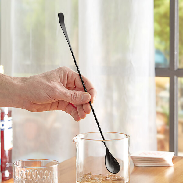A hand holding an Acopa black Japanese bar spoon in a glass of liquid.