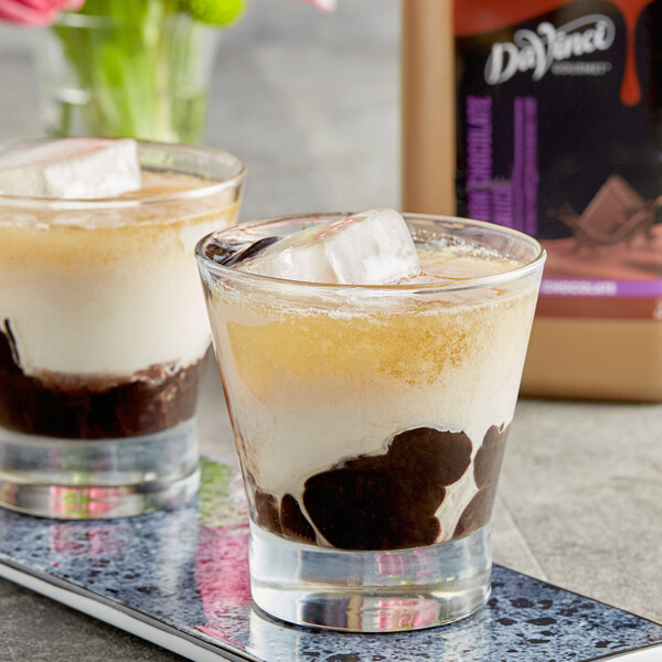 Two glasses of liquid with ice cubes and DaVinci Gourmet Dark Chocolate Flavoring Sauce on the side.