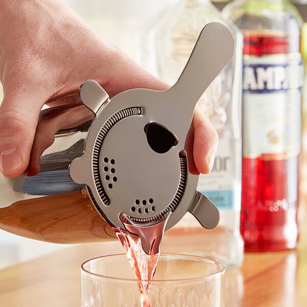 A hand using an Acopa 4 Prong Black Hawthorne Strainer to pour a drink from a metal container into a glass.