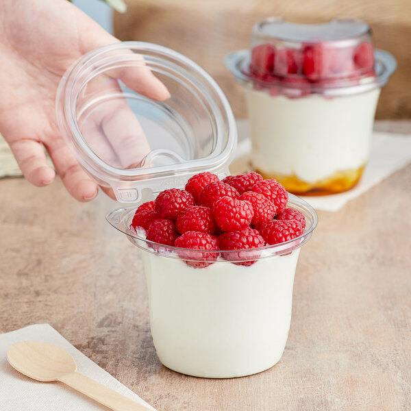 A hand holding a 12 oz. clear plastic cup of yogurt with raspberries and a wooden spoon.