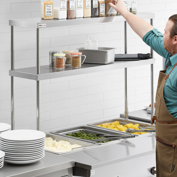 A man in an apron reaches for food on a double overshelf over a steam table.