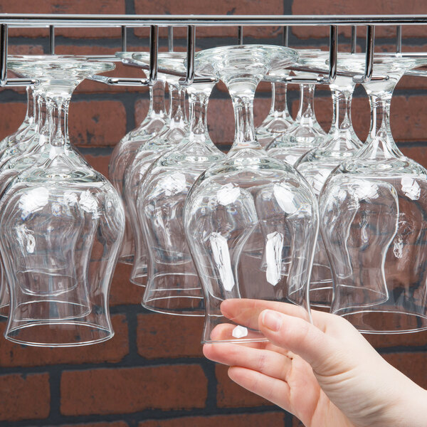 A hand holding a wine glass in front of a few wine glasses hanging on a Regency chrome wire bar glass rack.