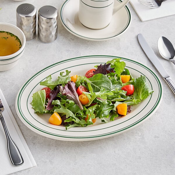 An Acopa ivory stoneware platter with green bands holding a salad with a fork on a white surface.
