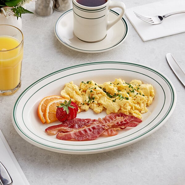 An Acopa ivory platter with green bands holding scrambled eggs, bacon, and fruit on a table.