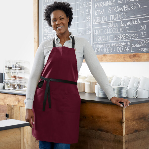 A smiling woman wearing a burgundy poly-cotton bib apron with black webbing accents.