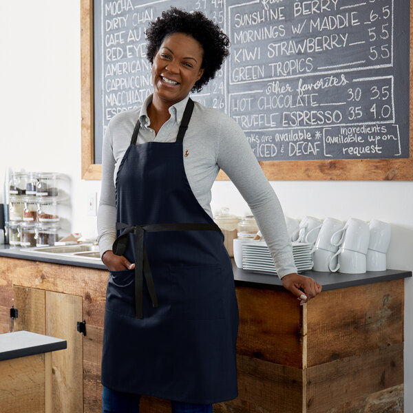 A woman wearing a navy blue bib apron with black accents stands in front of a counter.