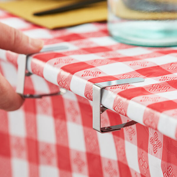 A hand using a Choice stainless steel tablecloth clip to secure a red and white checkered tablecloth on a table.