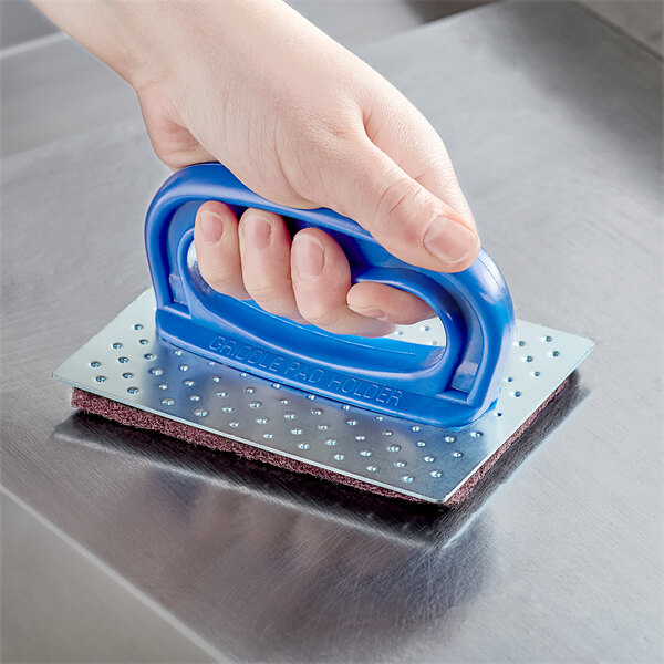 A hand with a blue handle using a Carlisle grill pad holder on a metal surface.