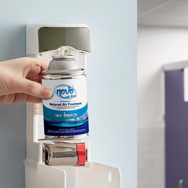 A hand holding a small silver can of Noble Chemical Sea Breeze air freshener in front of a wall mounted dispenser.