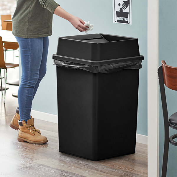 A woman standing next to a Lavex black square trash can with swing lid.
