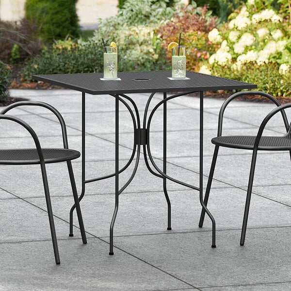 A Lancaster Table & Seating Harbor black metal table with ornate legs and two chairs on a stone patio.