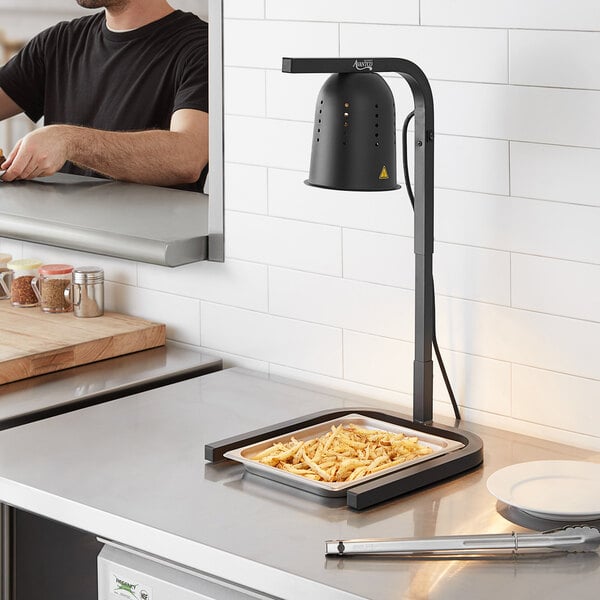 A man using an Avantco black free standing heat lamp to cook food on a countertop in a professional kitchen.