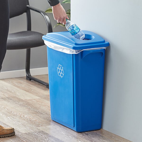 A woman's hand throws a plastic bottle into a blue Lavex Slim Rectangular recycle bin.