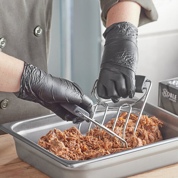 A person wearing black Noble disposable gloves stirs food in a metal pan on a counter.