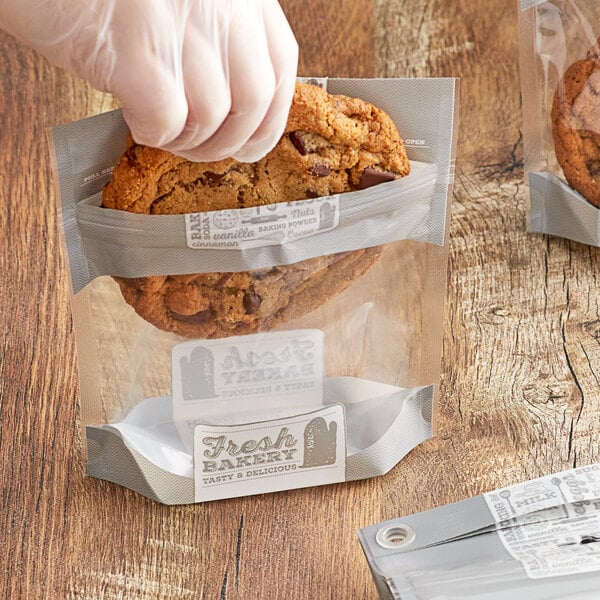 A gloved hand places a cookie in a small "Fresh Bakery" plastic bag.