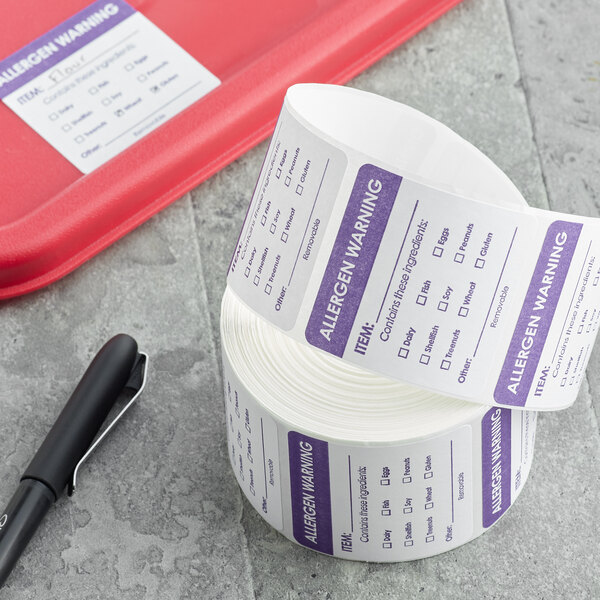 A roll of Noble Products allergen warning labels with white and purple writing next to a black pen on a grey surface.
