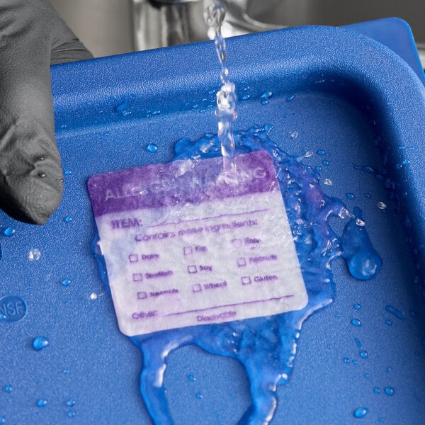 A dissolvable allergen label being rinsed off a blue food container under running water.