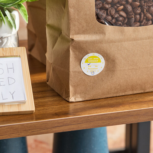A brown paper bag with a clear window showing coffee beans and a round yellow and white day-of-the-week clock label marked 'Tuesday' attached to the front.