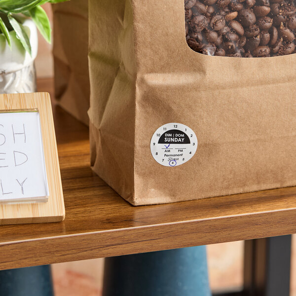 A brown paper bag with a clear window showing coffee beans and a round Sunday day-of-the-week clock label attached to the front.