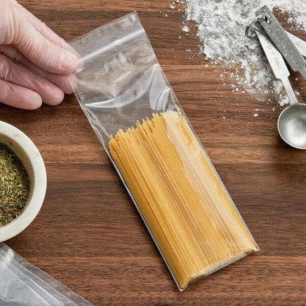A hand holding a Clear Line Seal Top Plastic Food Bag of pasta over a bowl of dried herbs.