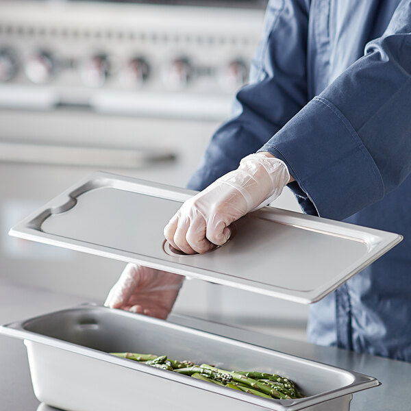 A person in a blue uniform holding a Vigor stainless steel long pan cover over a tray of food.